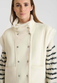 Ivoor fleecvest met een hoge kraag, zilveren knopen en voorkruiden. De mouwen hebben zwart-witte gestreepte bontaccenten. Zachte textuur.