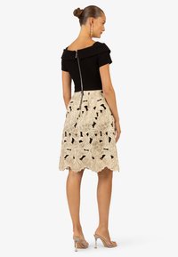 Top nero aderente con scollo off-the-shoulder, zip sulla schiena; gonna beige ricamata con ritagli floreali e orlo a onda.