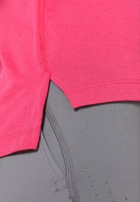 Rosa träningströja med strukturerad yta, med en delad nederkant, i kombination med grå tighta leggings som visar subtila prickmönster och sömnadsdetaljer.