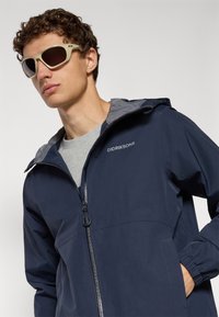 Jeune homme avec des cheveux bouclés portant des lunettes de soleil beiges et une veste à capuche navy Didriksons par-dessus une chemise grise, main dans la poche.