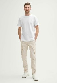 Weißes T-Shirt mit kurzen Ärmeln, kombiniert mit hellbeigen Cargo-Hosen mit Seitentaschen, getragen mit weißen Sneakern vor einem neutralen Hintergrund.