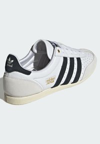 Tenisi albi adidas cu dungi negre, accente din piele întoarsă și un logo auriu. Au o textură perforată și o talpă din cauciuc texturată.