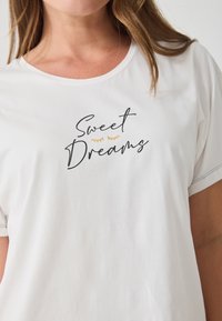 Fehér, rövid ujjú póló, puha anyagból készült, fekete és arany színű „Sweet Dreams” grafikával, amely alatt szempillák díszítik a szöveget.