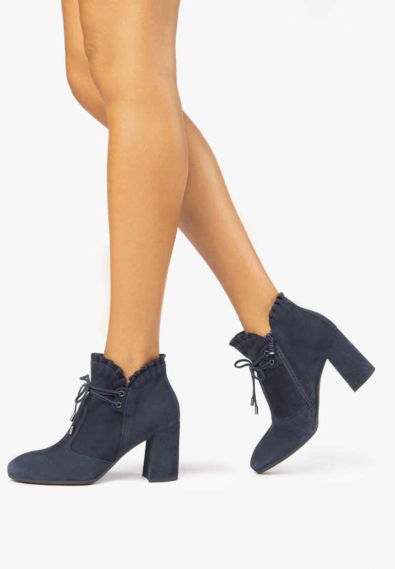 Botas de tobillo azul de ante con un alto tacón bloque, cuello con volantes y diseño con cordones. Textura suave y forma de punta redonda.