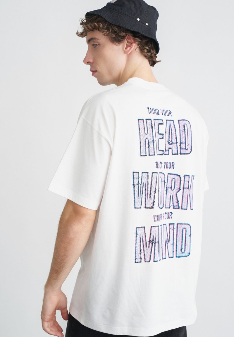 Biała bawełniana koszulka z krótkim rękawem, z wielokolorowym napisem na plecach: "MIND YOUR HEAD, WORK YOUR MIND", wyświetlanym pionowo.