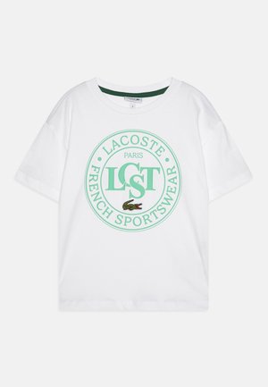 T-shirt bianco a maniche corte realizzato in cotone, con un logo circolare verde che presenta la scritta "LACOSTE PARIS FRENCH SPORTSWEAR" e un coccodrillo ricamato.