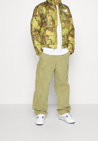 Nadýchaná zelená maskáčová bunda se žlutými akcenty přes bílou košili, doplněná světle zelenými nákladními kalhotami a bílými teniskami.