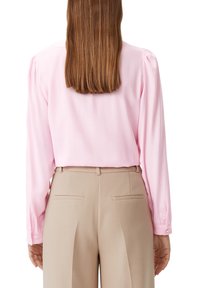 Blouse rose pâle à manches longues avec de subtils plis aux épaules, associée à un pantalon beige taille haute avec poches arrière et coupe ajustée.