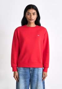 Red sweatshirt made of soft fabric, featuring a round neck, ribbed cuffs, and a small logo on the chest. Paired with light blue denim.