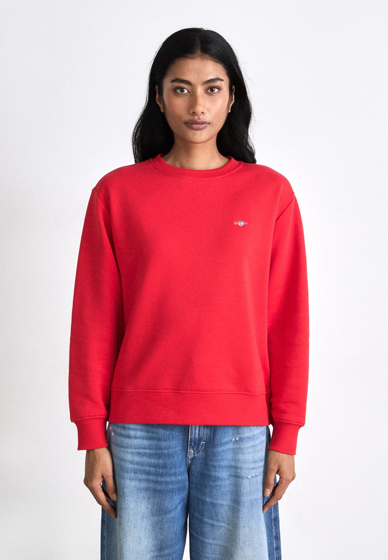 Red sweatshirt made of soft fabric, featuring a round neck, ribbed cuffs, and a small logo on the chest. Paired with light blue denim.