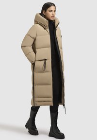 Lang, beige dunjakke med hætte, med lynlås foran, sidelommer og quiltet tekstur. Sammen med sorte kampstøvler.