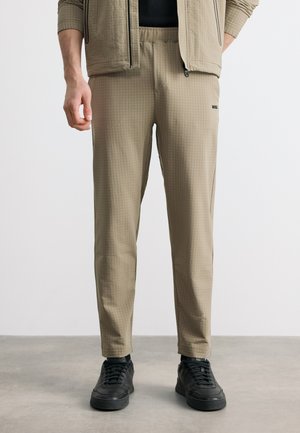 Muž oblečený v béžových textúrovaných úzkych nohaviciach, čiernych teniskách a béžovej bunde v rovnakom odtieni, stojaci na sivom podlaží s uvoľnenými rukami.