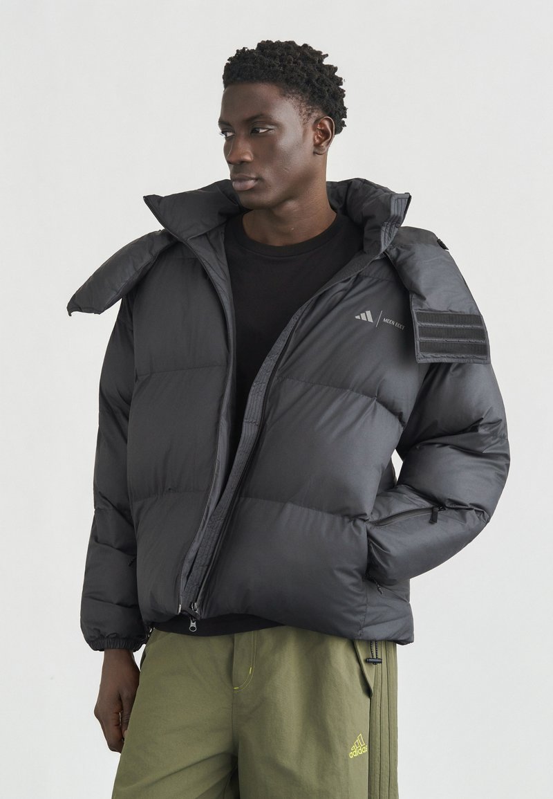 Νέος άνδρας που φοράει μαύρο μπουφάν puffer και χακί παντελόνι Adidas, κοιτάζοντας προς τα αριστερά του, μπροστά σε απλό φωτεινό φόντο.