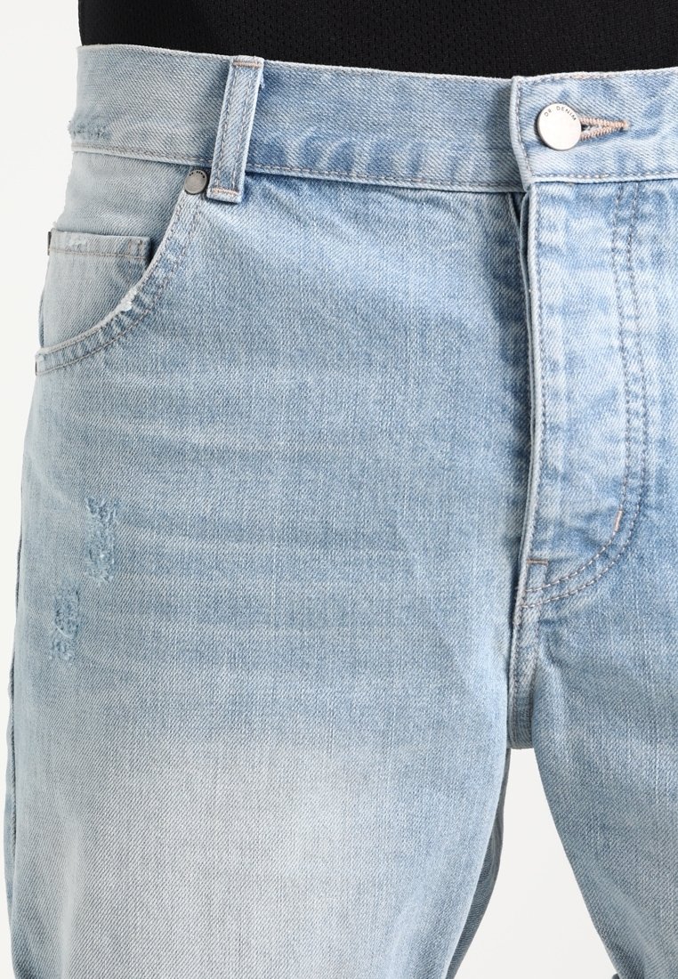 Jean en denim bleu clair, zone de la taille et du haut de la cuisse, avec un bouton à l'avant, des passants pour ceinture et une légère usure près de la poche.