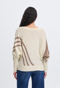 Maglione a maniche lunghe color crema con motivi a onda marroni, vestibilità rilassata e maniche ampie. Tessuto morbido, con vita e polsini a coste.