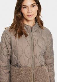 Vadderad beige jacka med ståkrage, hel dragkedja framtill och en texturerad brun fleecepanel nedtill. Slät, lättviktsmaterial.