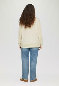 Beiger Strickpullover mit langen Ärmeln, lockerer Passform und geripptem Saum, kombiniert mit hellblauen geraden Jeans und Schuhen mit brauner Sohle.