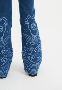Blå denim jeans med 3D broderede fisk og bølger designet langs benene, med en tekstureret overflade og en kraftig sort sål.