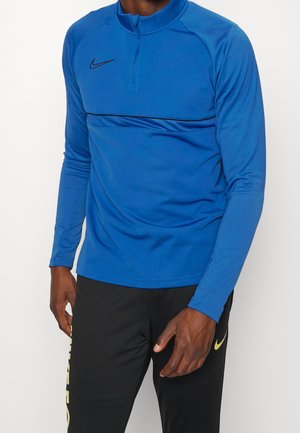 Mann trägt ein blaues langärmeliges Sportoberteil mit Frontreißverschluss und schwarze Hose mit gelbem Branding nahe dem Oberschenkel.