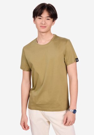 Junger Mann mit kurzen dunklen Haaren, der ein olivgrünes kurzärmeliges T-Shirt und helle Hosen vor weißem Hintergrund trägt.