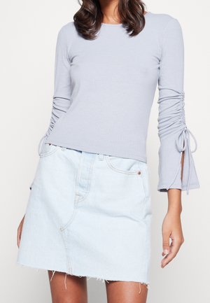 Femme portant un haut bleu clair à manches longues côtelées avec des manches froncées et une jupe trapèze en denim clair à ourlet brut.