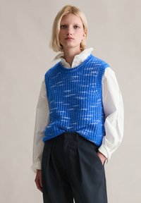 Niebieski dziergany kamizelka z teksturowanym wzorem i białymi akcentami, noszona na białej koszuli z guzikami i zestawiona z ciemnymi spodniami.