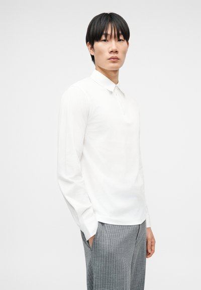 Νέος άνδρας με μαύρα μαλλιά, φοράει λευκό πουκάμισο μακρυμάνικο και γκρι παντελόνι με ρίγες, στέκεται με τα χέρια στις τσέπες μπροστά σε απλό φόντο.