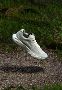 Light gray athletic shoe with a textured upper, rounded toe, and padded collar. Rubber sole with traction patterns. Floating above a gravel path.