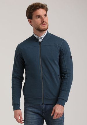 Zip-up jacket in teal fabric with a round collar, side pockets, and contrasting stitch details. Worn over a patterned shirt.