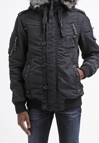 Giacca nera con cappuccio foderato in pelliccia, bottoni frontali, diverse tasche con cerniera, polsini a coste e una finitura texturizzata. Indossata con jeans blu.