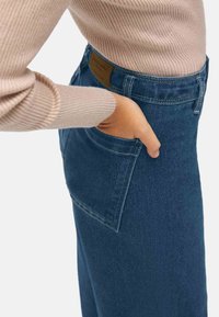 Vaqueros de mezclilla en tela azul oscuro con un corte ajustado, que cuentan con bolsillos traseros y costuras visibles. Acompañados de un suéter de canalé en un tono beige claro.