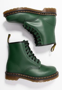 Botas de tobillo de cuero verde con cordones negros, punta redonda, costuras amarillas y una suela de goma con patrón. Incluyen una lengüeta en el talón.