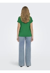 Femme aux longs cheveux blonds, vue de dos, portant une chemise verte à manches courtes et un jean bleu clair à jambes larges sur un fond uni.
