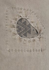 Patch de tissu beige avec une découpe en forme de goutte révélant un matériau à motif en dessous, cousu avec un fil contrastant. Détail texturé entourant la découpe.