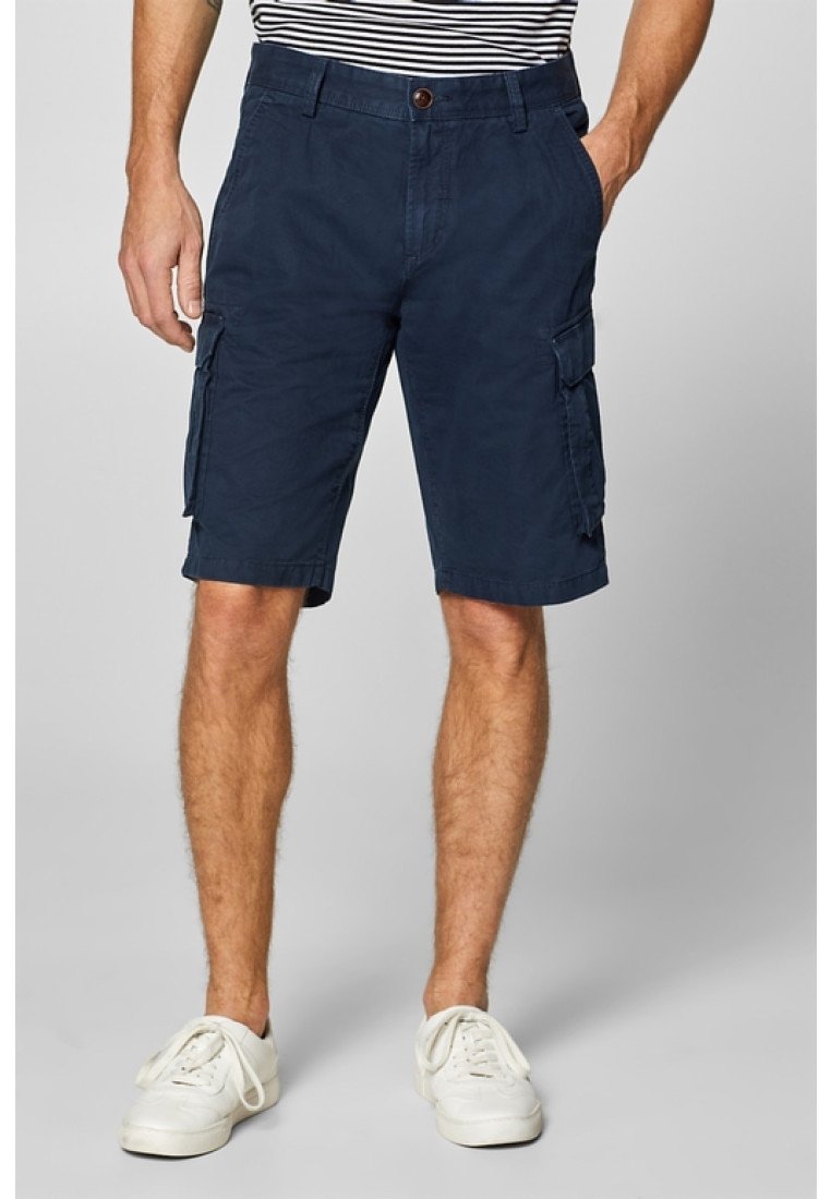 Homme portant un short cargo bleu marine, une chemise rayée rentrée et des baskets blanches, debout les mains dans les poches.