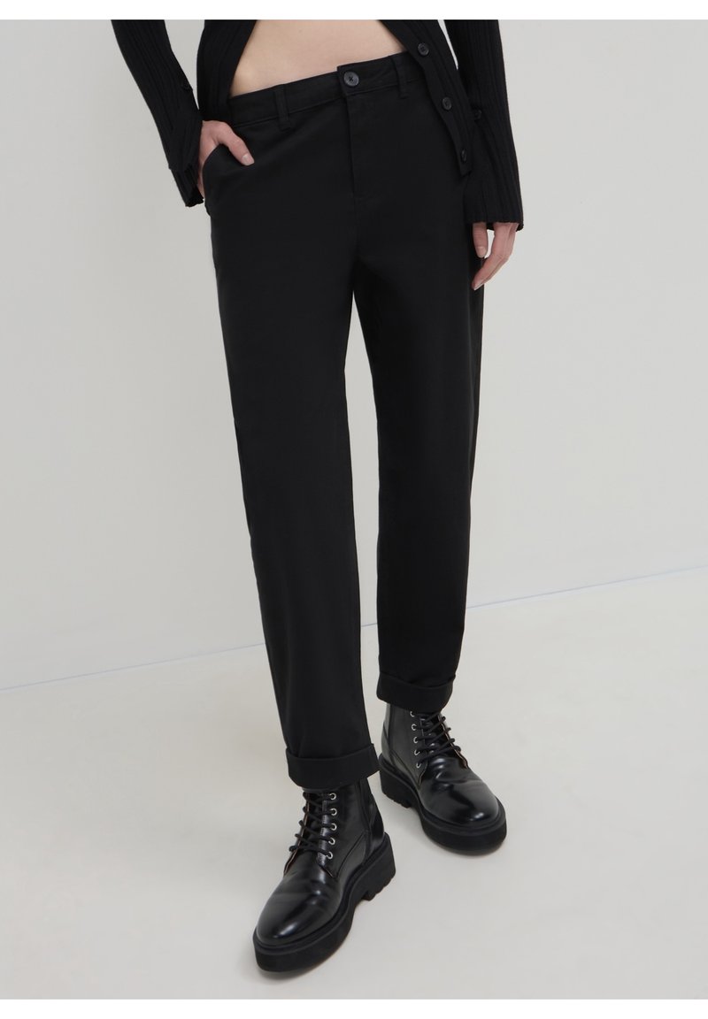 Personne portant un pantalon noir à revers, des bottes noires à lacets et un haut noir à manches longues, une main dans la poche.