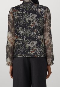 Vista posteriore di una persona che indossa una blusa trasparente nera con fiori e maniche lunghe, accompagnata da pantaloni grigio scuro.