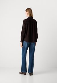 Woman wearing a dark long-sleeve shirt and blue jeans, standing facing away, on a plain white background.