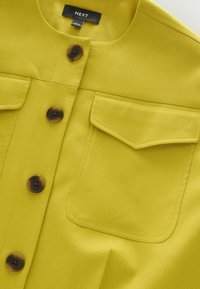 Žltozelená košeľa na gombíky so zaobleným golierom, štyrmi hnedými gombíkmi a dvoma náprsnými vreckami s klopou, označená nápisom „NEXT“, vyrobená v Bangladéši.