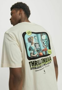 Lys cremefarvet t-shirt med et grafisk motiv på bagsiden, der indeholder vintage horror-elementer, herunder kranier, et spejl og farverige tekstdetaljer.