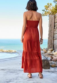 Robe maxi en orange rouille, avec un corsage smocké, une jupe à volants et une texture lisse. Portée avec des sandales marron clair. Arrière-plan avec vue sur la mer.