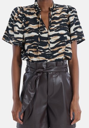 Vrouw draagt een blouse met korte mouwen in zwart, beige en wit dierenprint en hoge, donkere bruine leren shorts met een gebonden riem.