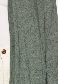 Zielony, teksturowany kardigan o luźnym kroju. Tkanina ma nakrapiany wzór. Posiada otwarty przód, zestawiony z kremowym topem.