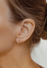 Pendiente de aro dorado que presenta una fila de pequeñas piedras transparentes a lo largo de un borde. El diseño es suave y circular, complementando la forma de la oreja.