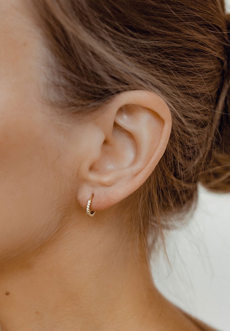 Pendiente de aro dorado que presenta una fila de pequeñas piedras transparentes a lo largo de un borde. El diseño es suave y circular, complementando la forma de la oreja.