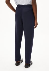 Pantaloni navy con vita elastica e texture liscia, dotati di tasca posteriore e gambe affusolate, abbinati a scarpe nere basse.
