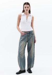Haut blanc sans manches avec un col zippé, associé à un jean baggy bleu clair présentant une texture délavée et de grandes poches. Des chaussures noires complètent le look.