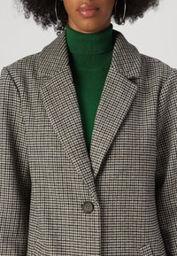 Blazer de cuadro de espiga en tonos terrenales, con cierre de botón marrón único, solapa en punta, superpuesto a un cuello de tortuga verde. Tejido texturizado.