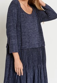 Marinblå, strukturerad långärmad topp med rundad hals, lagerdesign, ihop med en plisserad midikjol. Mjuk, avslappnad tyg.