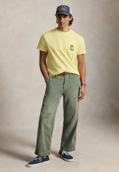 Νεαρός άνδρας που φοράει κίτρινο t-shirt με σχέδιο φοίνικα, πράσινο φαρδύ παντελόνι, μπλε αθλητικά παπούτσια και μπλε καπέλο Polo Team, στέκεται χαλαρός.